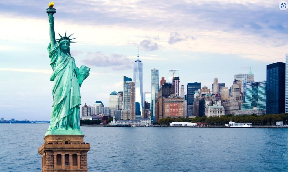 The Statue of Liberty with its iconic torch held high in the foreground, and the New York City skyline featuring the One World Trade Center in the background under a cloudy sky.